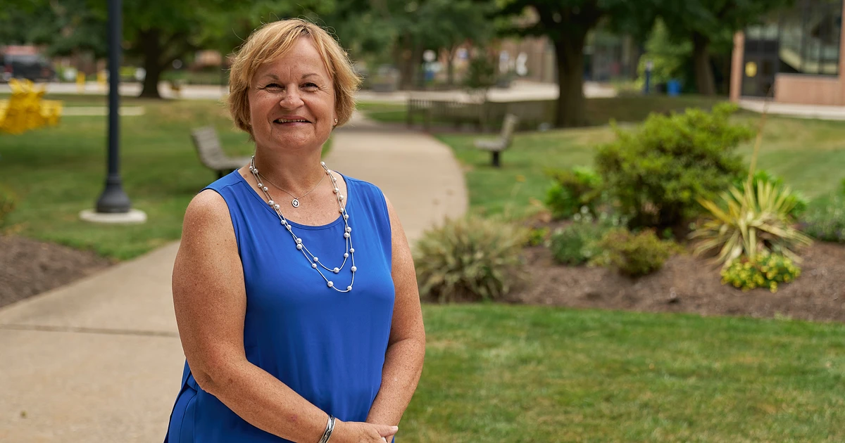 A woman in business casual clothing, a blue tank top, smiling outside in front of greenery