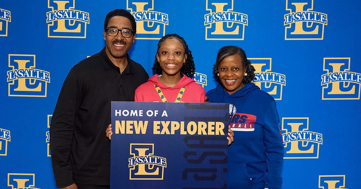 Three people pose happily in front of a La Salle backdrop holding a sign that reads "Home of a New Explorer."